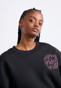 Čierna mikina so zaobleným fialovým logom s textom "alpha industries." Má pohodlný, voľný strih a rebrovaný výstrih.