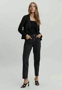 Crni slim fit blejzer, crni gornji dio, crne traperice visokog struka i crne sandale s visokom petom nosi osoba s dugom kosom. Glatke teksture, elegantan dizajn.