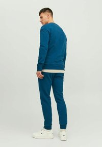 Felpa blu e jogger abbinati realizzati in tessuto morbido; presentano polsini a costine, design aderente e sneakers bianche. Vista posteriore.