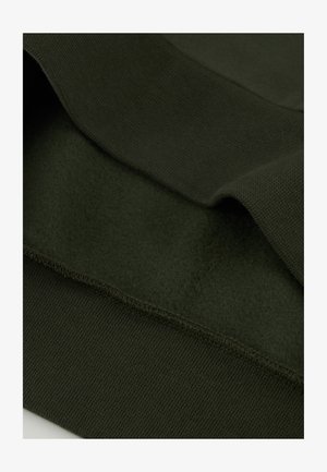 Ciemnozielony polar o gładkiej fakturze, z ryflem w pasie i na końcach для zapewnienia dopasowania. Warstwowy wygląd podkreśla miękkość.