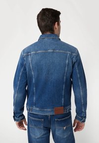 Denimjas in medium blauw met een klassieke snit, voorzien van een omklapbare kraag, lange mouwen en een leren merkpatch op de achterkant.