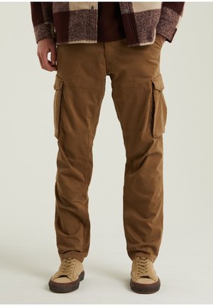 Mężczyzna w brązowych spodniach typu cargo z bocznymi kieszeniami, beżowych tenisówkach i bordowo-beżowej kratkowanej kurtce, stojący na jednolitym tle.