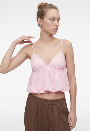 Młoda kobieta poprawia ramiączko luźnej różowej bezrękawnika, nosi brązowe spodnie z wysokim stanem, na jednolitym białym tle.