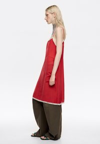 Blondynka w profilu nosi czerwoną sukienkę na ramiączkach na luźnych oliwkowych spodniach i turkusowych sandałach, uzupełnioną długim srebrnym naszyjnikiem typu choker.