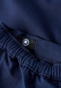 Marineblått stoff med elastisk midjebånd og justerbar svart stropp festet med en klar plastknapp.