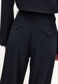 Pantalon noir au design ajusté, avec deux poches arrière à rabats pointus. Le tissu présente une texture lisse et une taille cintrée.