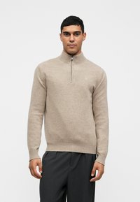 Bēšas krāsas džemperis ar rāvējslēdzēja apkakli, izgatavots no maiga materiāla. Tam ir garas piedurknes un ribots odere un apakša. Minimālistisks dizains ar tīru logo detaļu.