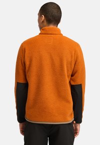 Orange Fleecejacke mit hohem Kragen, strukturierter Oberfläche und schwarzen Stoffakzenten an den Ärmeln. Mit einem verstellbaren Saum.