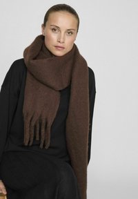 Écharpe en laine marron avec des franges, enroulée autour du cou, assortie à un haut noir. Texture douce, épaisseur considérable.