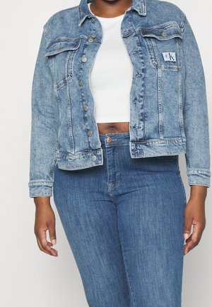 Person iført en lyseblå denimjakke over en hvid crop top og matchende blå højttaljede jeans, stående foran en ensfarvet baggrund.