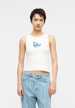 Γυναίκα που φοράει λευκό αμάνικο crop top με μπλε και πράσινο γραφικό και ανοιχτόχρωμα τζιν με ρίγες, στέκεται μπροστά σε απλό φόντο.