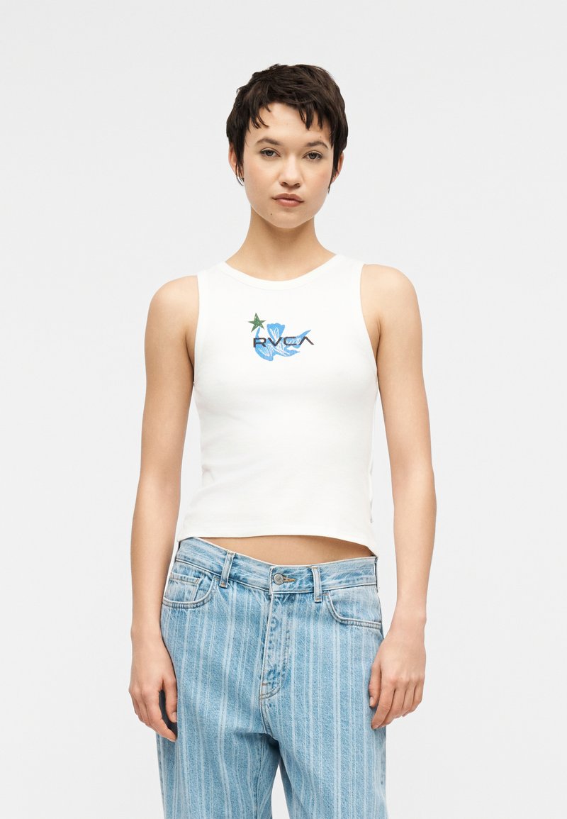 Γυναίκα που φοράει λευκό αμάνικο crop top με μπλε και πράσινο γραφικό και ανοιχτόχρωμα τζιν με ρίγες, στέκεται μπροστά σε απλό φόντο.