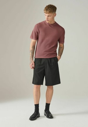 Young man with tattoos wearing a textured short-sleeve mauve shirt, black pleated shorts, black socks, and black loafers, standing and looking down.