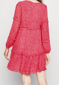 Donna che indossa un vestito rosso corto a maniche lunghe con stampa leopardata rosa e dettagli a volant, vista da dietro su uno sfondo semplice.