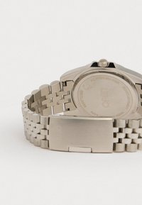 Hopeakromitettu ruostumaton teräksinen kello, jossa on teksturoitu ranneke ja pyöreä kotelo. Takapuolella on kaiverrettua tekstiä ja kruunu sivussa.