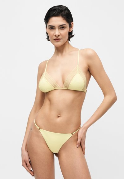 Γυναίκα με μπικίνι ανοιχτού κίτρινου χρώματος με δαντέλα, στέκεται με το ένα χέρι στη μέση σε απλό ανοιχτό φόντο.