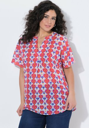 Vrouw met krullend haar, gekleed in een kleurrijk, geometrisch patroon korte mouwen blouse en blauwe jeans, staand tegen een witte achtergrond.