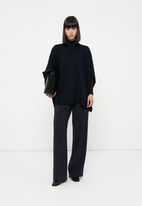 Maglione poncho di colore navy in un morbido filato, caratterizzato da un collo alto e maniche larghe. Abbinato a pantaloni scuri, larghi e a una pochette nera.