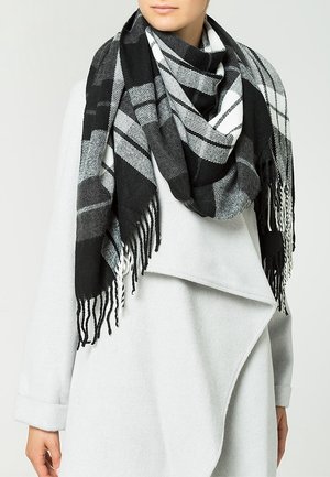 Femme portant un manteau gris clair avec une grande écharpe à carreaux noire, blanche et grise enroulée autour du cou, aux extrémités frangées.