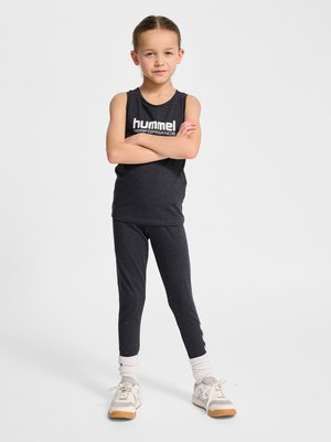 Jong meisje staat zelfverzekerd met gekruiste armen, draagt een zwarte mouwloze top, bijpassende legging, witte sokken en beige sneakers.