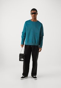 Teal hettegenser, svarte jeans og svarte joggesko. Holder en svart bag med hvit logo. Solbriller fullfører looken.