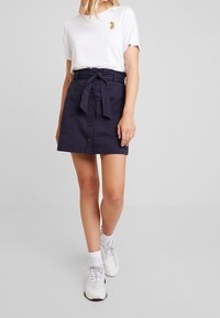 Gonna in cotone blu navy con cintura a fusciacca, bottoni frontali e tasche. Abbinata a una t-shirt bianca con un piccolo logo colorato.