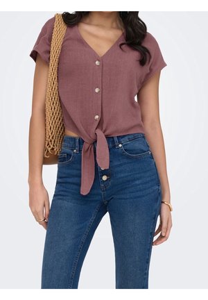 Vrouw draagt een mauve gekleurde, kortärmelig top met knopen, gestrikt in de taille, blauwe hooggesneden spijkerbroek, een geweven schoudertas en een gouden armband.