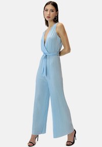 Jumpsuit plisado azul claro con escote en V profundo, cintura ceñida con cinturón y piernas anchas. Tejido suave con un brillo sutil, complementado por tacones altos negros.