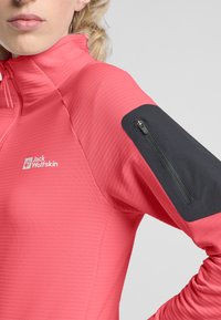 Rosa Zip-Jacke aus strukturiertem Material, mit einer schwarzen Reißverschlusstasche auf der Schulter und einem Logo auf der Brust.