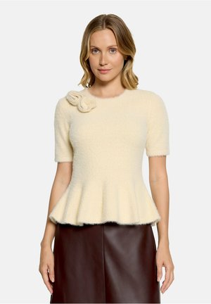 Femme portant un haut duveteux blanc crème à manches courtes avec un ourlet peplum et un détail décoratif de fleur près de l'encolure, associé à une jupe foncée.