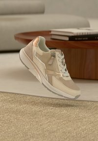 Μπεζ αθλητικό παπούτσι με υλικά από δίχτυ και σουέντ, λευκά κορδόνια, μεταλλικές λεπτομέρειες και υφή στη σόλα, εμφανιζόμενο πάνω σε ουδέτερο φόντο.