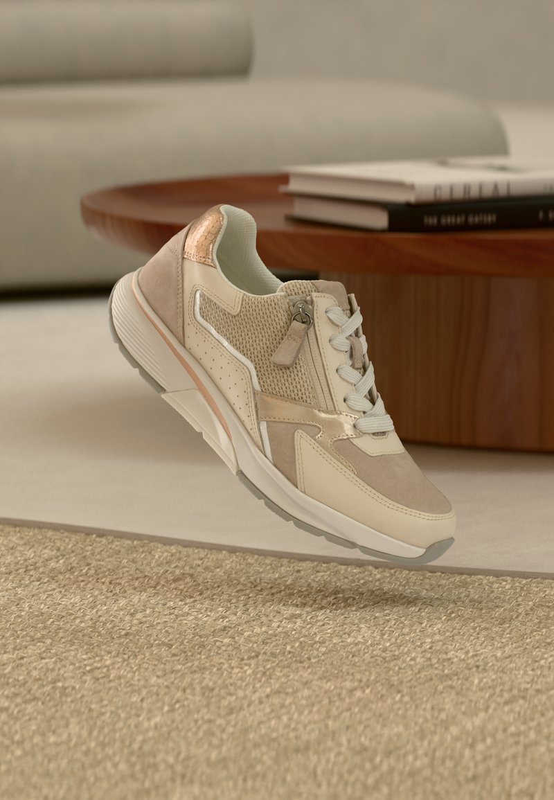 Μπεζ αθλητικό παπούτσι με υλικά από δίχτυ και σουέντ, λευκά κορδόνια, μεταλλικές λεπτομέρειες και υφή στη σόλα, εμφανιζόμενο πάνω σε ουδέτερο φόντο.