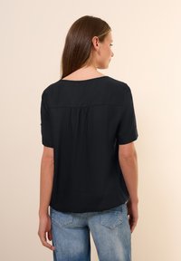 Sort kortærmet top med afslappet pasform, featuring en bådudskæring, rynket detalje bagpå og et struktureret stof. Kombineret med blå jeans.
