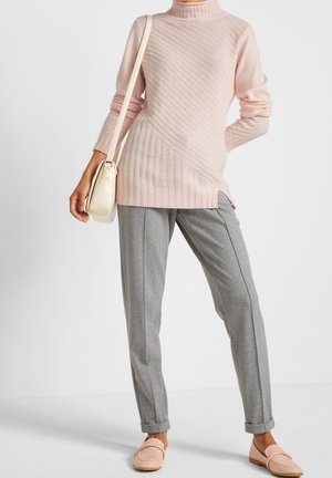 Femme portant un pull à col roulé rose clair, un pantalon gris ajusté, des mocassins roses et un petit sac à épaule beige.