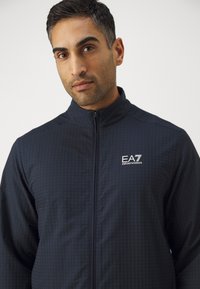 Marineblauwe zip-up jack met een gewatteerd patroon, voorzien van een hoge kraag en het EA7-logo op de linkerbogen. Glad, lichtgewicht stof.