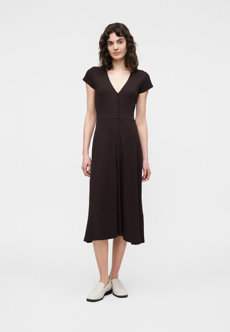 Mujer de pie con un vestido midi negro de manga corta y botones, escote en V y zapatos blancos sin cordones sobre un fondo blanco.