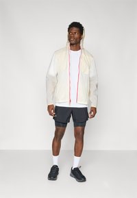 Giacca leggera e semitrasparente beige con cappuccio, dotata di cordini rossi. Indossata sopra una camicia bianca e shorts neri, con sneaker nere.