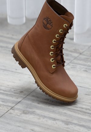 Braune Leder Schnürstiefel mit dicker Sohle und eingeprägtem Baum-Logo, auf einem hellgrauen Fliesenboden in der Nähe eines weißen vertikalen Paneels platziert.