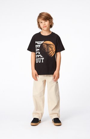 T-shirt nera con grafica di basket bianca e arancione e testo "PEACE OUT", abbinata a pantaloni beige chiari a gamba larga e sneakers nere.