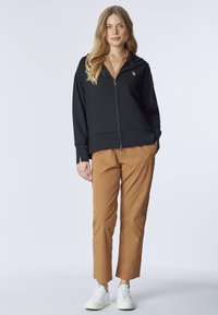 Felpa nera con zip e tasche frontali, indossata sopra una maglia marrone, abbinata a pantaloni beige e sneaker bianche. Tessuto morbido, vestibilità rilassata.