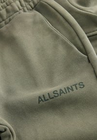 Pantaloni in misto cotone verde oliva con tasca visibile e il nome del marchio "ALLSAINTS" stampato in verde scuro sul davanti. Texture morbida.