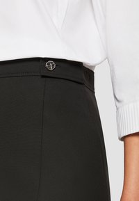 Musta housut, joissa on sileä pinta, hopeinen nappisuljenta ja korkeavyötäröinen malli. Vyötärönauha on siististi räätälöity.