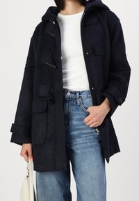 Persona vistiendo un abrigo de toggles azul marino oscuro sobre una camisa de canalé blanca y jeans azules, sosteniendo un bolso blanco con la mano izquierda.