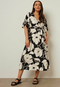 Svart omlottklänning med stora vita blommönster, korta ärmar och ett knytbälte. Bärs med svarta sandaler, som har platt sula och spännedetalj.