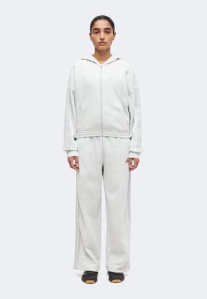 Persona de pie con chaqueta con capucha y cremallera gris claro y pantalones anchos a juego con zapatillas negras sobre fondo liso.