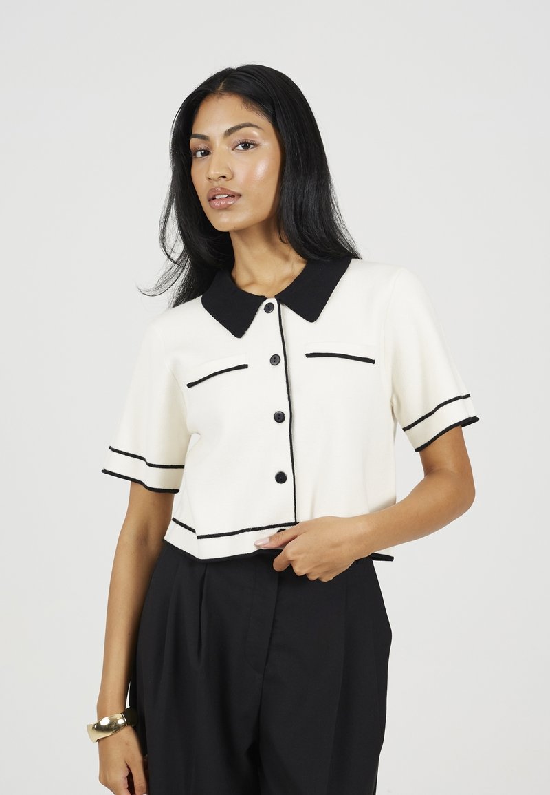 Cardigan blanc raccourci avec un col noir et des accents, doté de manches courtes, d'une fermeture à boutons et de deux poches avant soulignées de noir.