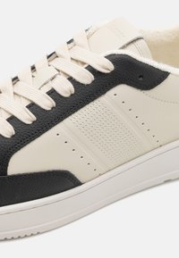 Baskets noires et crème avec un dessus en cuir texturé, des panneaux de couleurs contrastantes, des perforations et des lacets plats.