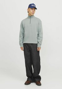 Light gray, zip-neck sweatshirt with long sleeves paired with loose black denim pants and brown shoes. Blue cap with text accents.