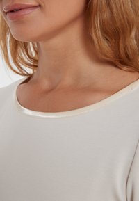 Lichtbeige top met een ronde halslijn, voorzien van een satijnen rand. De stof lijkt glad en zacht, en sluit zich voorzichtig rond het nekgebied.
