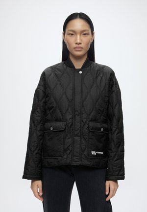 Ung kvinde står med fronten mod kameraet iført en sort quiltet jakke med trykknaplommer og et mærke med "Karl Lagerfeld Jeans" foran.